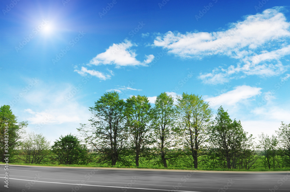 An empty countryside road in motion with trees and bushes against a blue sky with clouds