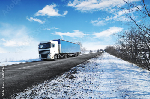 Truck with a trailer on the winter countryside road with snow against sky with clouds