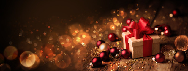 Abstract Christmas Banner  -  Gift box with red bow and baubles on wood  -  Magic dark golden bokeh lights background