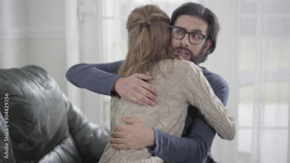 Young blond Caucasian woman showing her boyfriend or husband affirmative pregnancy test. Bearded man imitating joy and hugging girl. Shocked guy making upset face behind wife's back.