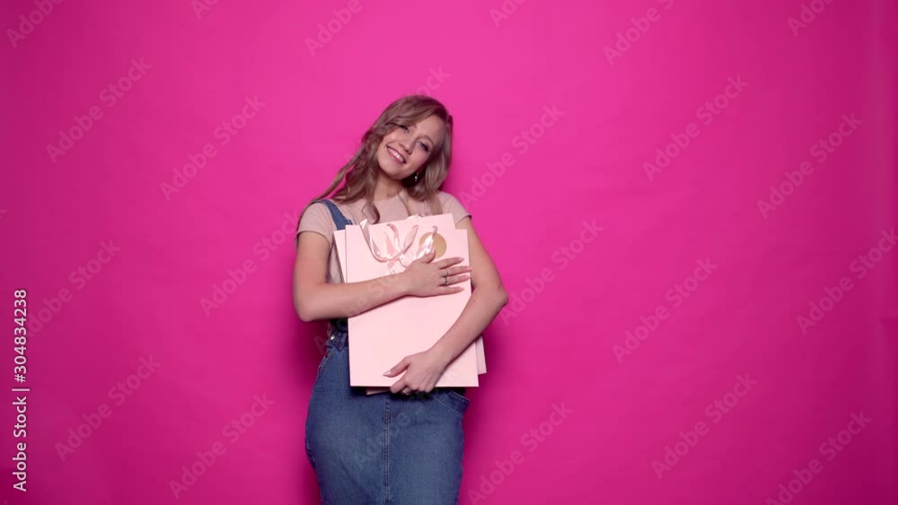 Happy young woman hugging her shopping bags, smiling and looking at the camera on isolated pink background. Cheerful girl in denim clothes posing with packages on season sale in professional studio. Happy young woman hugging her shopping bags, smiling and looking at the camera on isolated pink background. Cheerful girl in denim clothes posing with packages on season sale in professional studio.