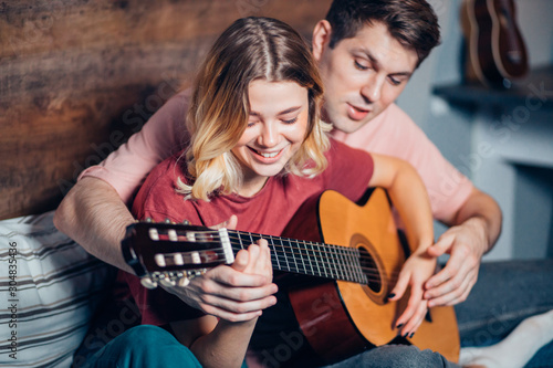 young boyfriend teaching her girlfriend to play the guitar at home, lovely couple wearing casual clothes in bedroom together