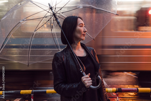 走っている電車の側に立つ、ビニール傘を指す女性のポートレート