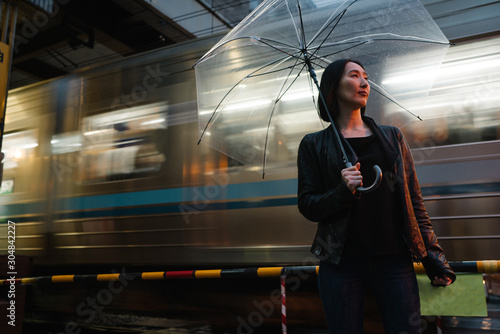 走っている電車の側に立つ、ビニール傘を指す女性のポートレート