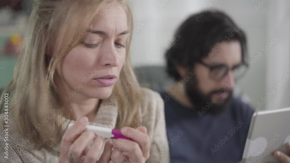 Young blond Caucasian girl with brown eyes holding pregnancy test and looking back at man using tablet at the background. Young woman getting ready to tell husband about pregnancy. Young blond Caucasian girl with brown eyes holding pregnancy test and looking back at man using tablet at the background. Young woman getting ready to tell husband about pregnancy.