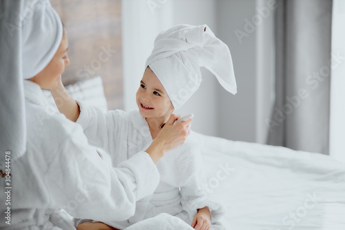 young caucasian woman sit in towel and bathrobe with daughter hold makeup brushes  white bedroom