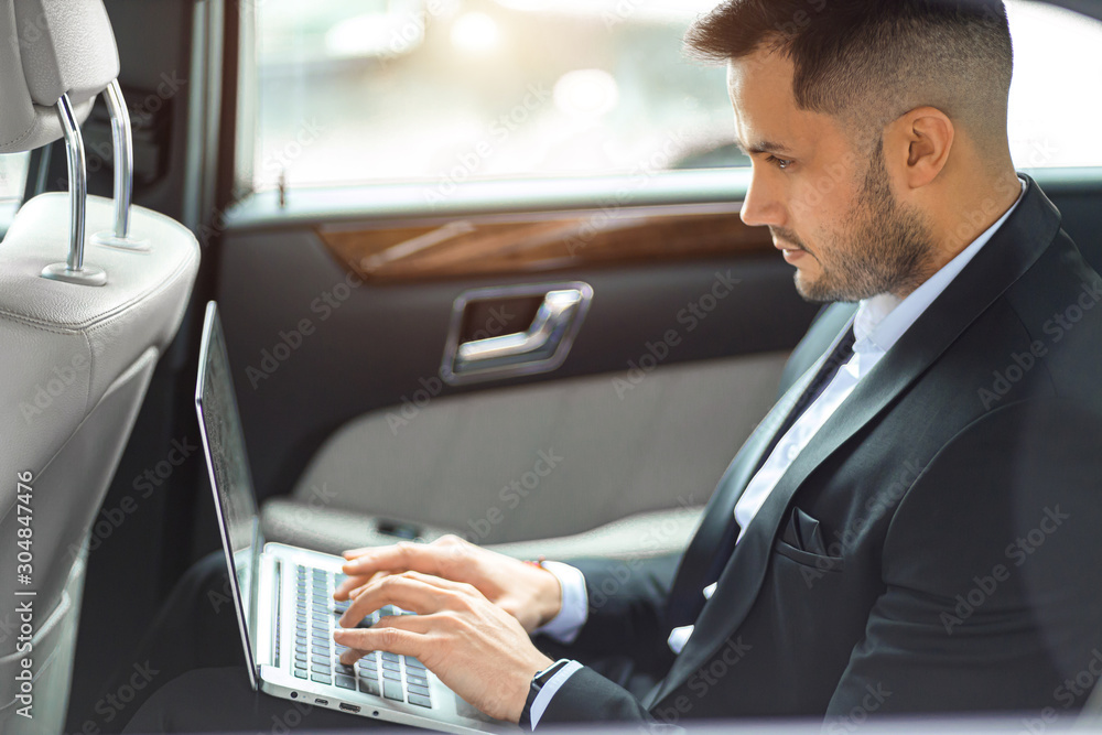 Caucasian man wearing formal wear sit with laptop in expensive luxurious car, side view