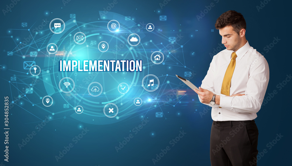 Businessman thinking in front of technology related icons and IMPLEMENTATION inscription, modern technology concept Businessman thinking in front of technology related icons and IMPLEMENTATION inscription, modern technology concept