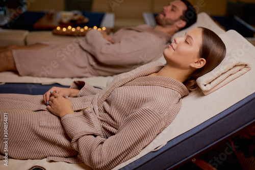 Young caucasian couple take rest in spa salon lying in bathrobes together and relax with candles