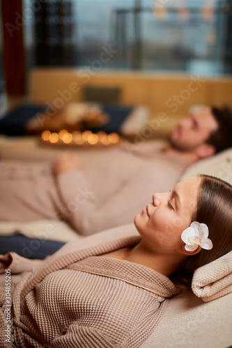 Young caucasian couple take rest in spa salon lying in bathrobes together and relax with candles