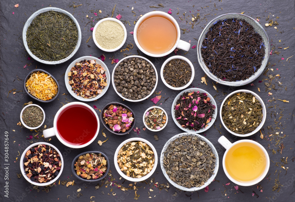 Assortment of tea cups, dried leaves, fruit and herbs in bowls on dark, stone background, top view Assortment of tea cups, dried leaves, fruit and herbs in bowls on dark, stone background, top view