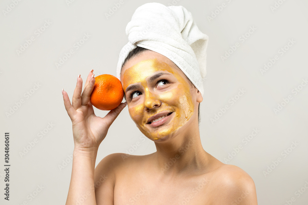 Beauty portrait of woman in white towel on head with gold nourishing mask on face. Skincare cleansing eco organic cosmetic spa relax concept. A girl stands with her back holding an orange mandarin. Beauty portrait of woman in white towel on head with gold nourishing mask on face. Skincare cleansing eco organic cosmetic spa relax concept. A girl stands with her back holding an orange mandarin.