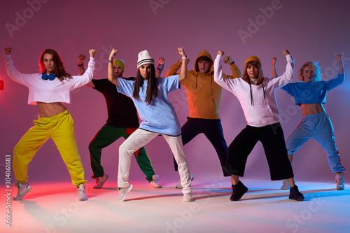Group of energetic street dancers in sportswear, standing on brightly lighted stage, raising hands up, keeping legs wide, performing dance movie, moving synchronically