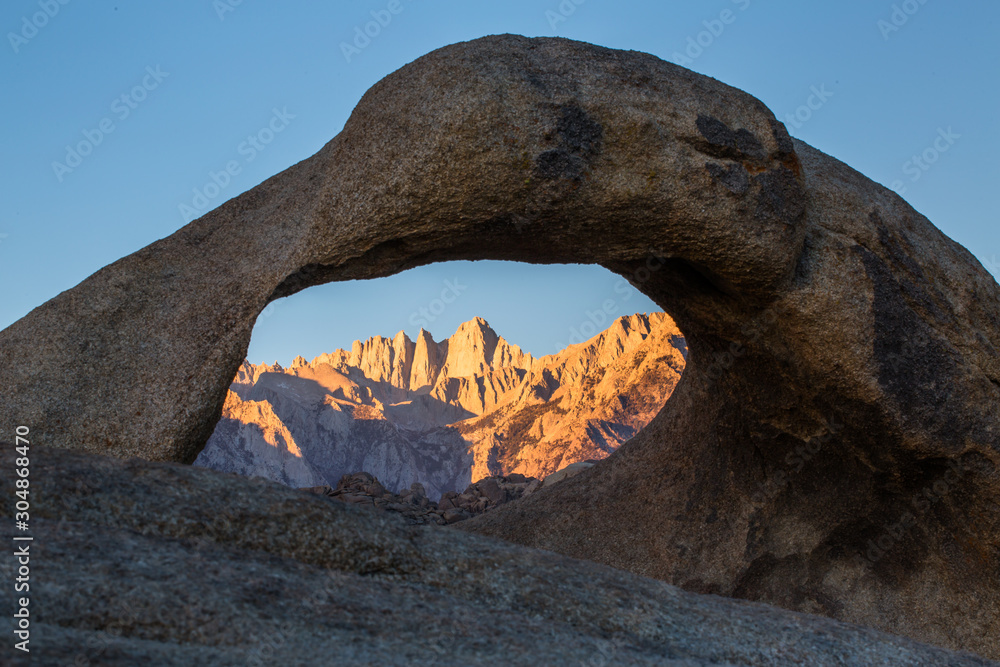 Mount Whitney in sunrise light through arch with clear sky overhead.