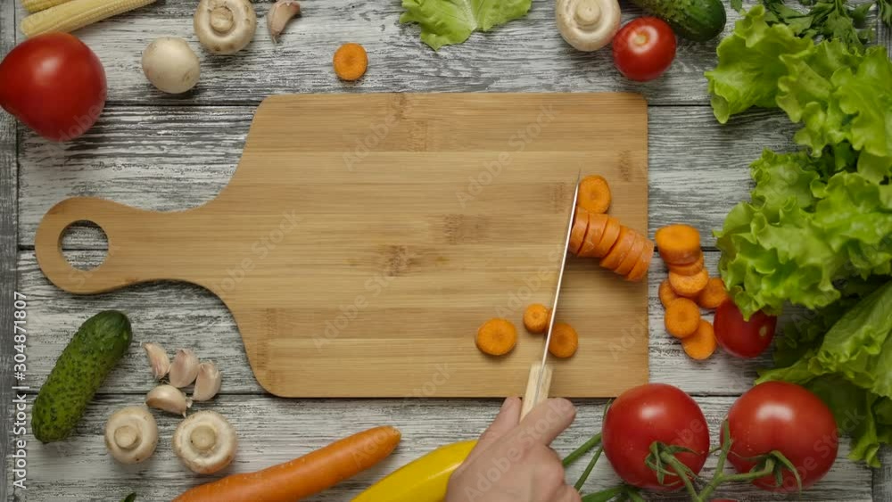 From above of cook pushing with knife sliced carrot on wooden cutting board. Overhead shot of vegetables on kitchen table. Tomatoes, salad, mushrooms, cucumbers.