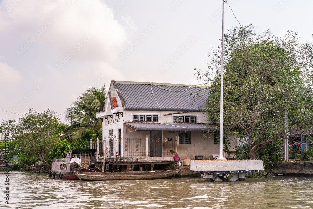 Cai Be, Mekong Delta, Vietnam - March 13, 2019: Along Kinh 28 canal. White high warehouse with gray roof on stilts under gray cloudscape and surrounded by green foliage. barge docked in front on brown