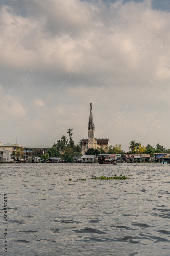 Cai Be, Mekong Delta, Vietnam - March 13, 2019: Along Kinh 28 canal. Portrait of local Catholic church behind row of retail businesses and green foliage under blue cloudscape. Dark water up front.