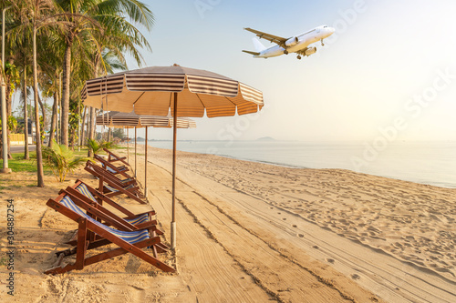 Row of empty wooden beach chairs and parasols on tropical sandy beach in the morning  with white passenger airplane landing above the sea , destinations relax summer holidays concept          