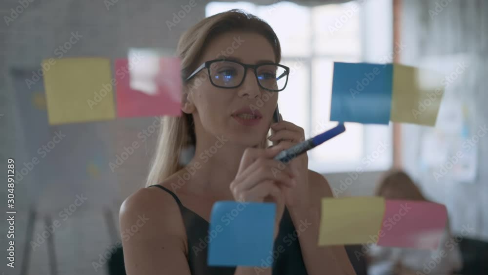 portrait of a successful girl medium shot, serious business woman talking on the phone and taking notes on stickers. modern office with glass wall 4K portrait of a successful girl medium shot, serious business woman talking on the phone and taking notes on stickers. modern office with glass wall 4K
