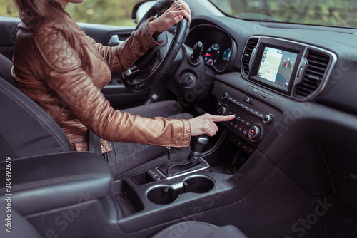 woman car interior includes button, application air conditioning selection, seat heating ventilation, automatic transmission, autumn spring forest park. Leather jacket. Screen with navigation.