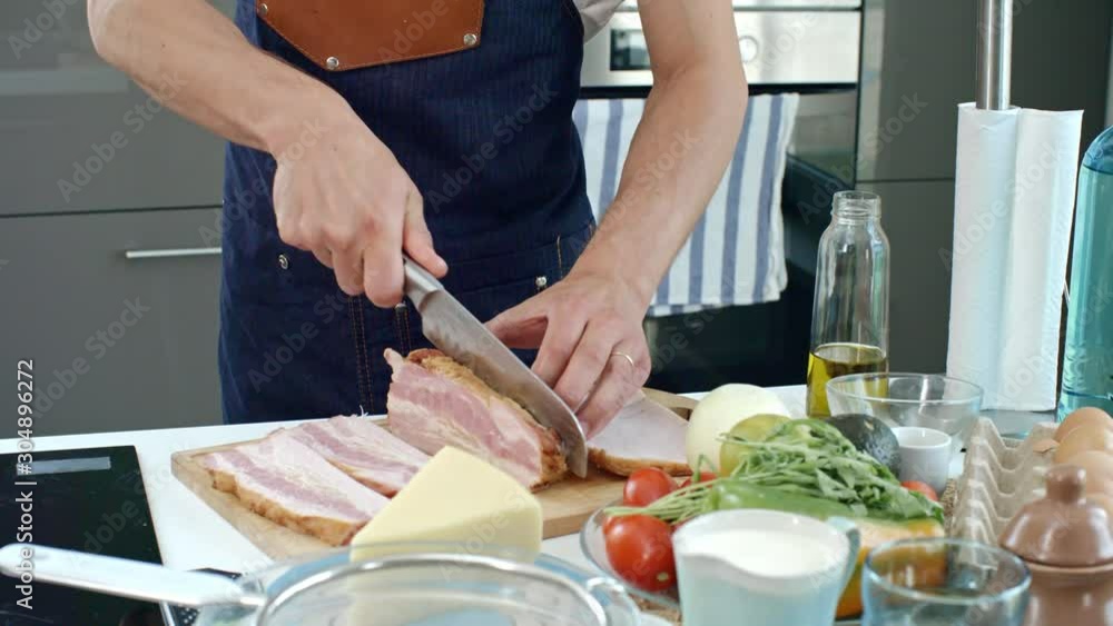 Unrecognizable male chef slicing bacon with sharp knife on wooden board while cooking in kitchen; cheese and fresh vegetables lying on table Unrecognizable male chef slicing bacon with sharp knife on wooden board while cooking in kitchen; cheese and fresh vegetables lying on table