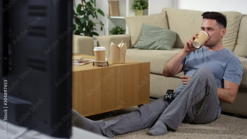 Cheerful middle aged man sitting on the floor in the living room, drinking coffee from disposal cup and changing channels with remote control while watching TV at home Cheerful middle aged man sitting on the floor in the living room, drinking coffee from disposal cup and changing channels with remote control while watching TV at home