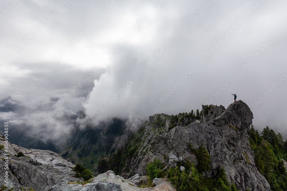 Adventurous Man Standing on top of a rugged rocky mountain during a cloudy summer morning. Taken on Crown Mountain, North Vancouver, BC, Canada.