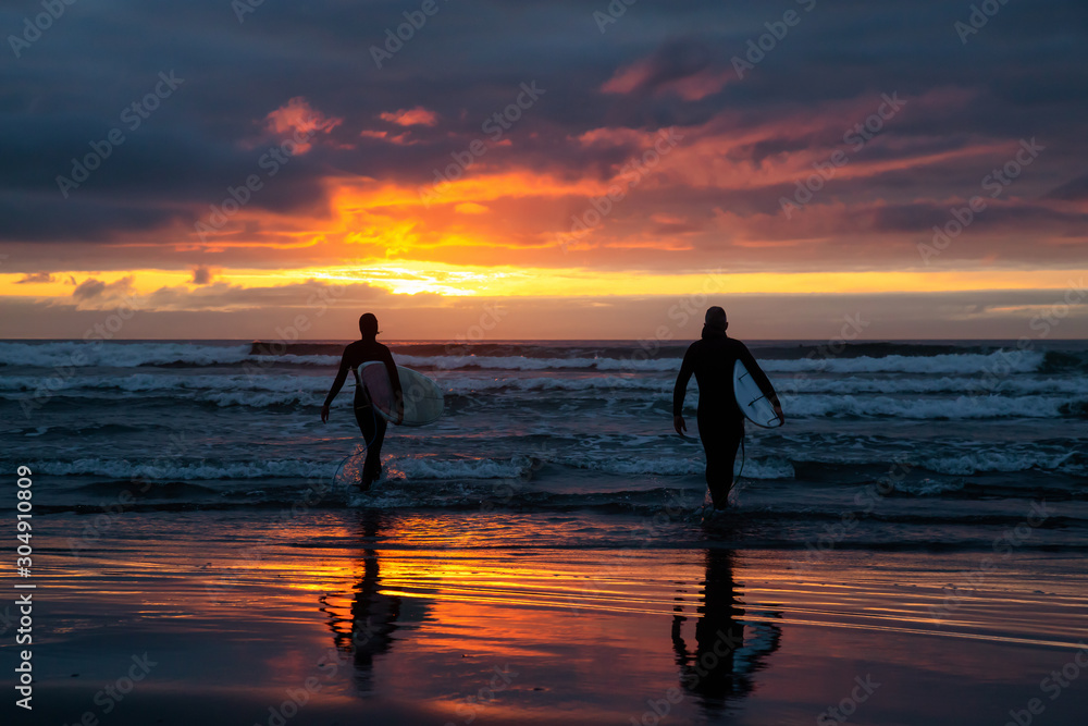 Surfers walking towards the Ocean on the sandy beach during a dramatic summer sunset. Taken in Seaside, Oregon, United States of America.