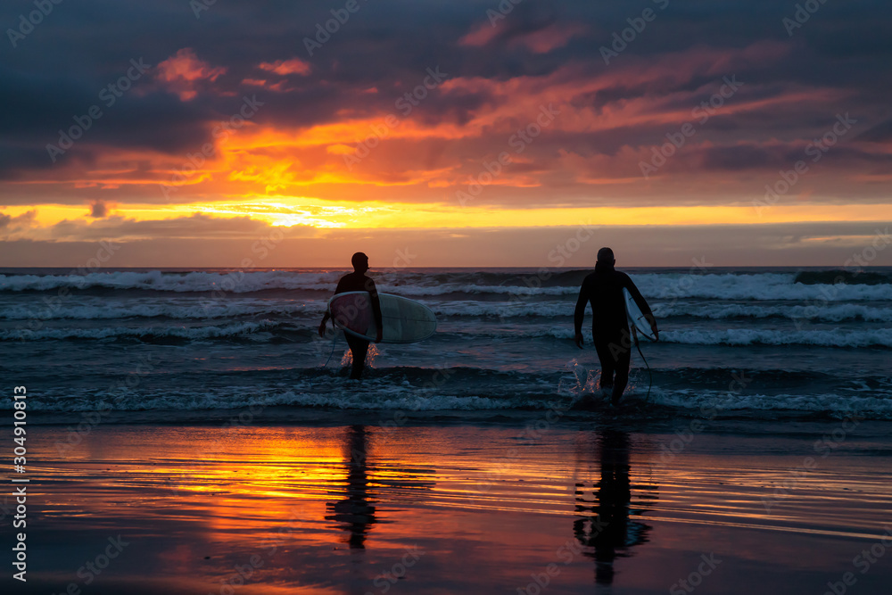 Surfers walking towards the Ocean on the sandy beach during a dramatic summer sunset. Taken in Seaside, Oregon, United States of America.