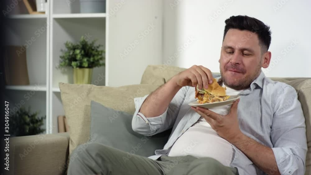Middle aged Caucasian man holding plate and eating delicious burger while sitting on couch and watching interesting program on TV in the living room Middle aged Caucasian man holding plate and eating delicious burger while sitting on couch and watching interesting program on TV in the living room