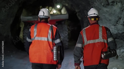 Two workers with helmets and equipment walking through a mine tunnel, in a background of heavy mining machinery.