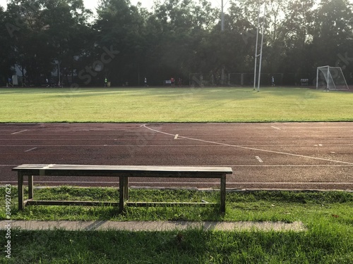 A wooden bench besides red rubber running track near the football field in the morning