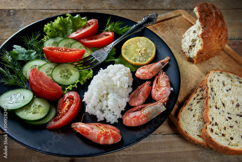Shrimp with rice and vegetables (tomatoes, cucumbers, lemon, dill and parsley) on a black plate. The bread on the board. 