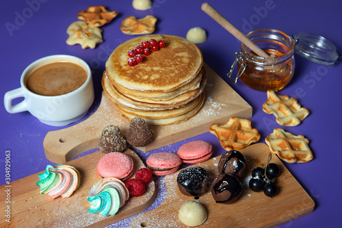 Pancakes with honey and sweets near to a cup of coffee on a purple background.