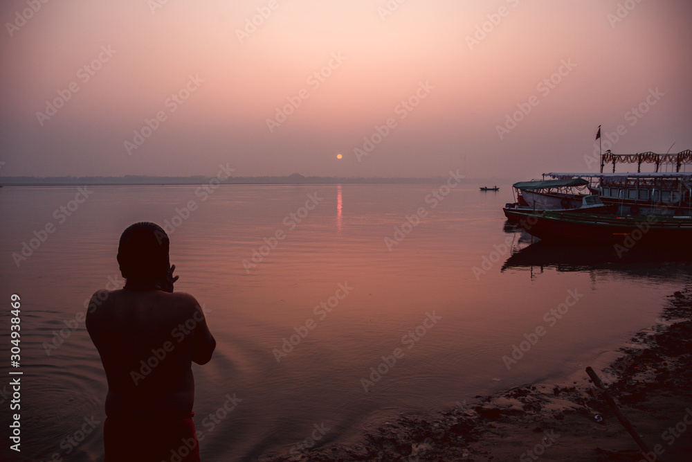 A hindu man praying during sunrise on the river Ganges in Varanasi, India. Fog, seagulls and boat silhouettes.