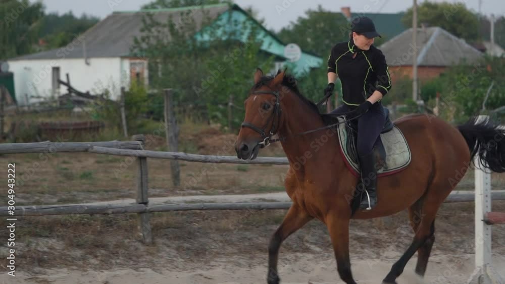 Horsewoman riding on brown horse and jumping the fence in sandy parkour riding arena. Competitive rider training jumping over obstacles in manege. Slow motion Horsewoman riding on brown horse and jumping the fence in sandy parkour riding arena. Competitive rider training jumping over obstacles in manege. Slow motion