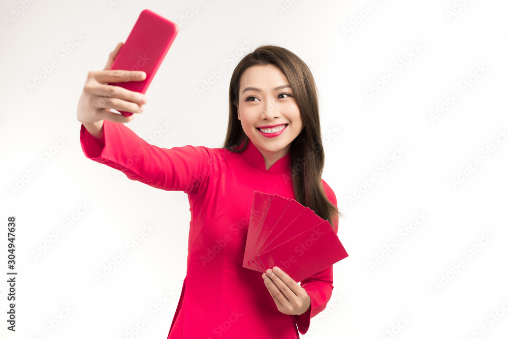 Portrait of a happy young girl dressed in ao dai taking a selfie while showing red envelope isolated over white background Portrait of a happy young girl dressed in ao dai taking a selfie while showing red envelope isolated over white background
