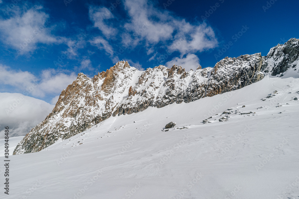 High alpine ridge in winter conditions. Rocky mountain ridge with ice and snow. Winter alpine landscape of Mount Blanc Massif, France. Blue sky and sunny day in high mountains.