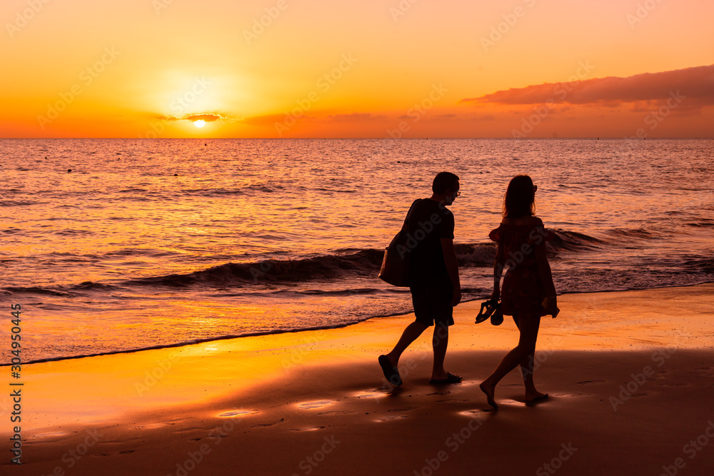 A man and a woman walk along the seashore against the backdrop of the setting sun. Orange sunset at the sea. Silhouettes on a sunset background