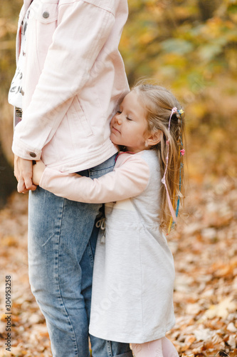 Smiling little girl standing with closed eyes and embracing her mother legs in the autumn parklegs in the autumn park