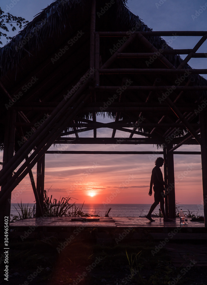 Man with sunset doing yoga. Tourist Man sitting and looking at beautiful orange, yellow and blue sunset over Montanita beach town from bamboo hut. Shot in Ecuador.