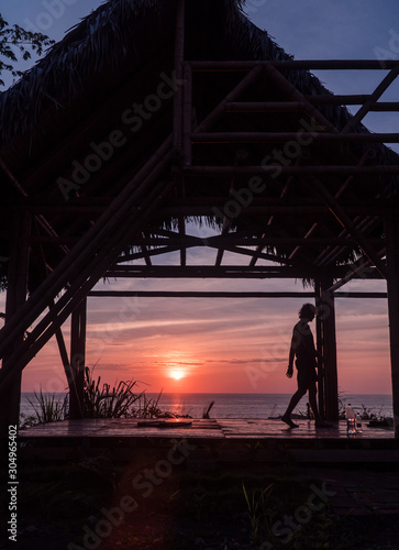 Man with sunset doing yoga. Tourist Man sitting and looking at beautiful orange, yellow and blue sunset over Montanita beach town from bamboo hut. Shot in Ecuador.