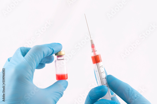 Doctor's hands in blue gloves holding syringe and bottle with red liquid on a white background
