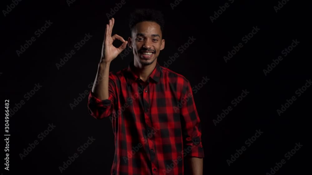 Handsome man in red wear on black studio background smiles to camera and gives ok sign. Happy african american guy showing gesture of approval. Winner.Success. Handsome man in red wear on black studio background smiles to camera and gives ok sign. Happy african american guy showing gesture of approval. Winner.Success.