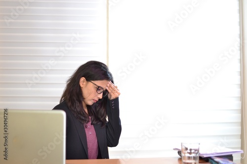 An office girl, smart, sweet and sexy is wearing black dress sitting on her desk and looking uncofortable, rubbing her hand over her forehead photo