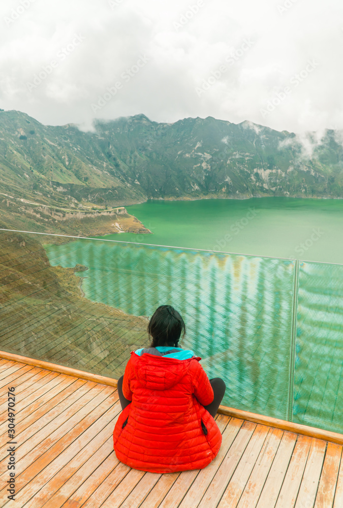 Fototapeta premium Woman at mountain volcano lake. Hiker in red looking at dramatic perspective of Quilotoa lake and crater view. Hiking loop from viewpoint. Shot in Ecuador. Green and blue. Freedom, Adventure.
