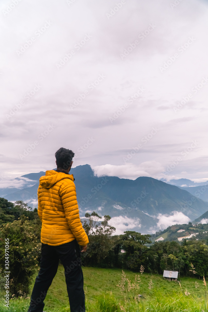 Hiker man looking at green mountains and cloudy sky, from viewpoint. Wearing yellow jacket, on holiday in Banos, Ecuador.