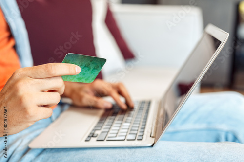 Leisure at Home. Young man sitting on sofa shopping onlien on laptop holding credit card close-up photo
