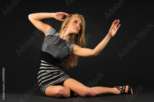 Young blond beautiful woman in black dress and shoes sitting on floor over dark background in photo studio. Beauty and fashion lifestyle concept