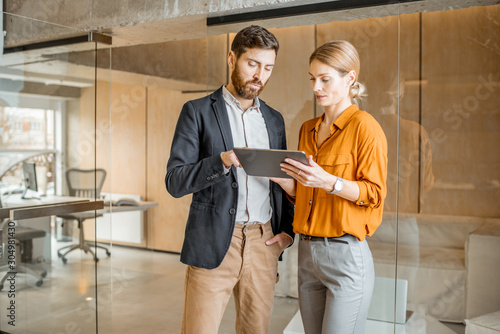 Two colleagues dressed casually meeting in the hallway, solving some working moments with digital tablet. Work in marketing agency or design studio concept